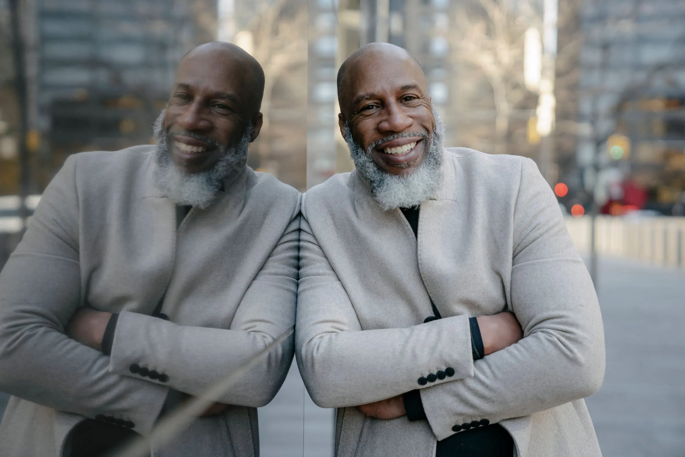 Smiling older man with a white beard leaning against a glass wall, arms folded